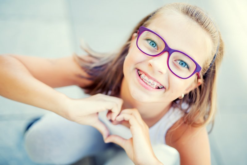 a young girl wearing purple glasses and braces makes a heart with her hands 